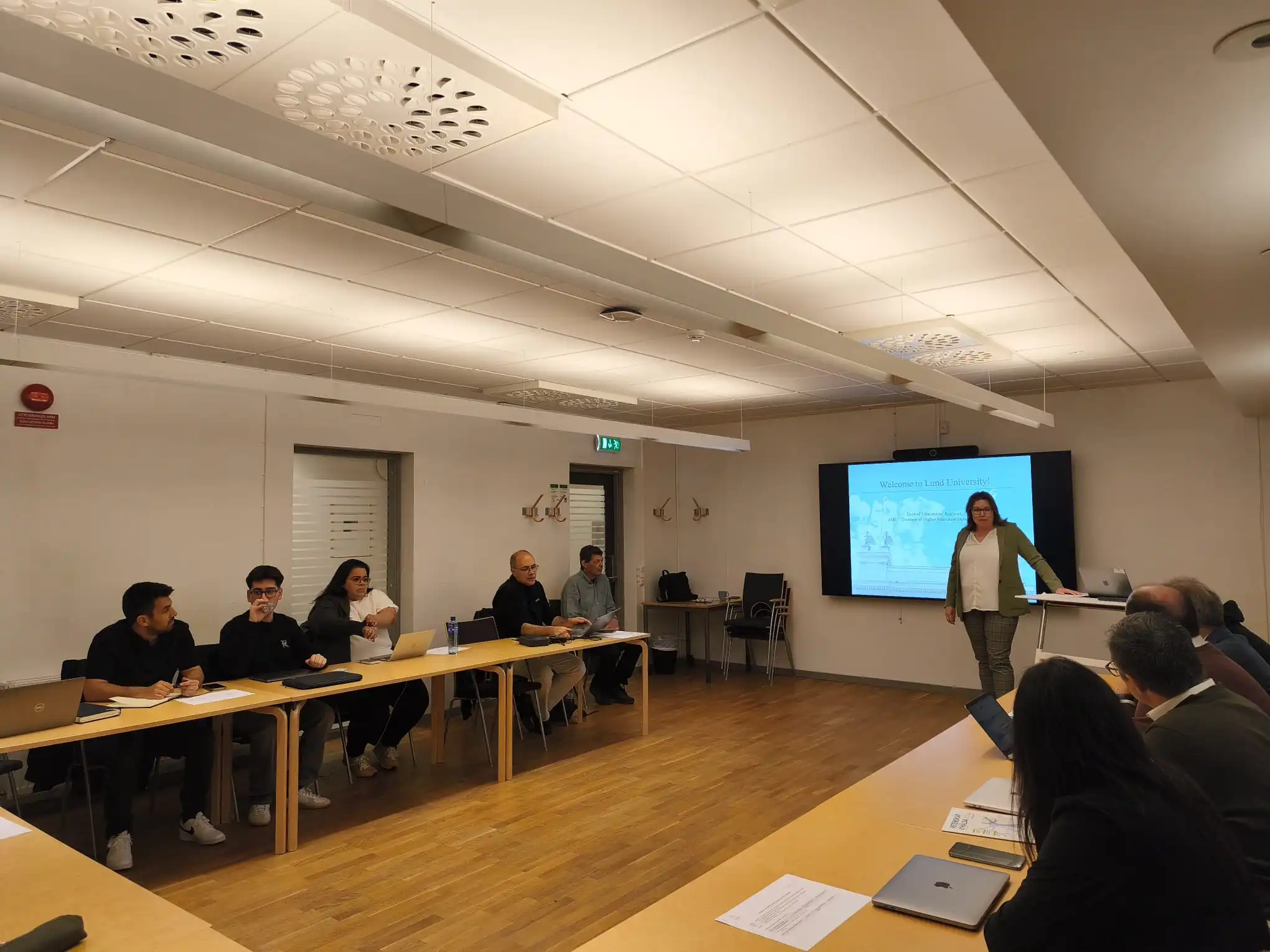 Reunião em sala com participantes sentados à volta de uma mesa e apresentação projetada ao fundo.