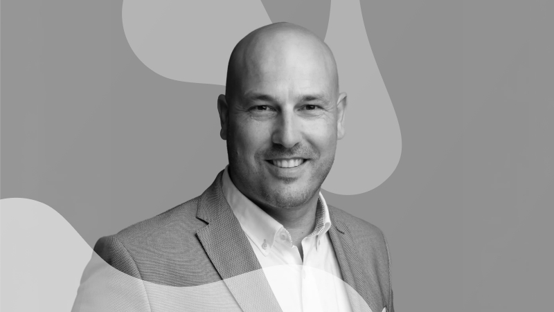 Retrato a preto e branco de um homem careca, de blazer e camisa, a sorrir sobre fundo gráfico abstrato.