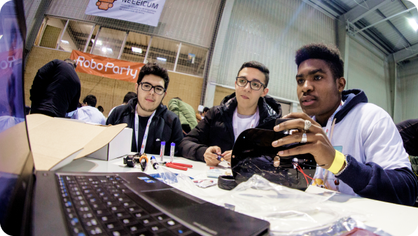 Grupo de participantes em contexto de aprendizagem prática, com equipamento tecnológico sobre a mesa.